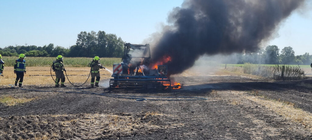 Einsatzkräfte löschen einen brennenden Traktor auf einem Stoppelfeld, dunkler Rauch steigt auf